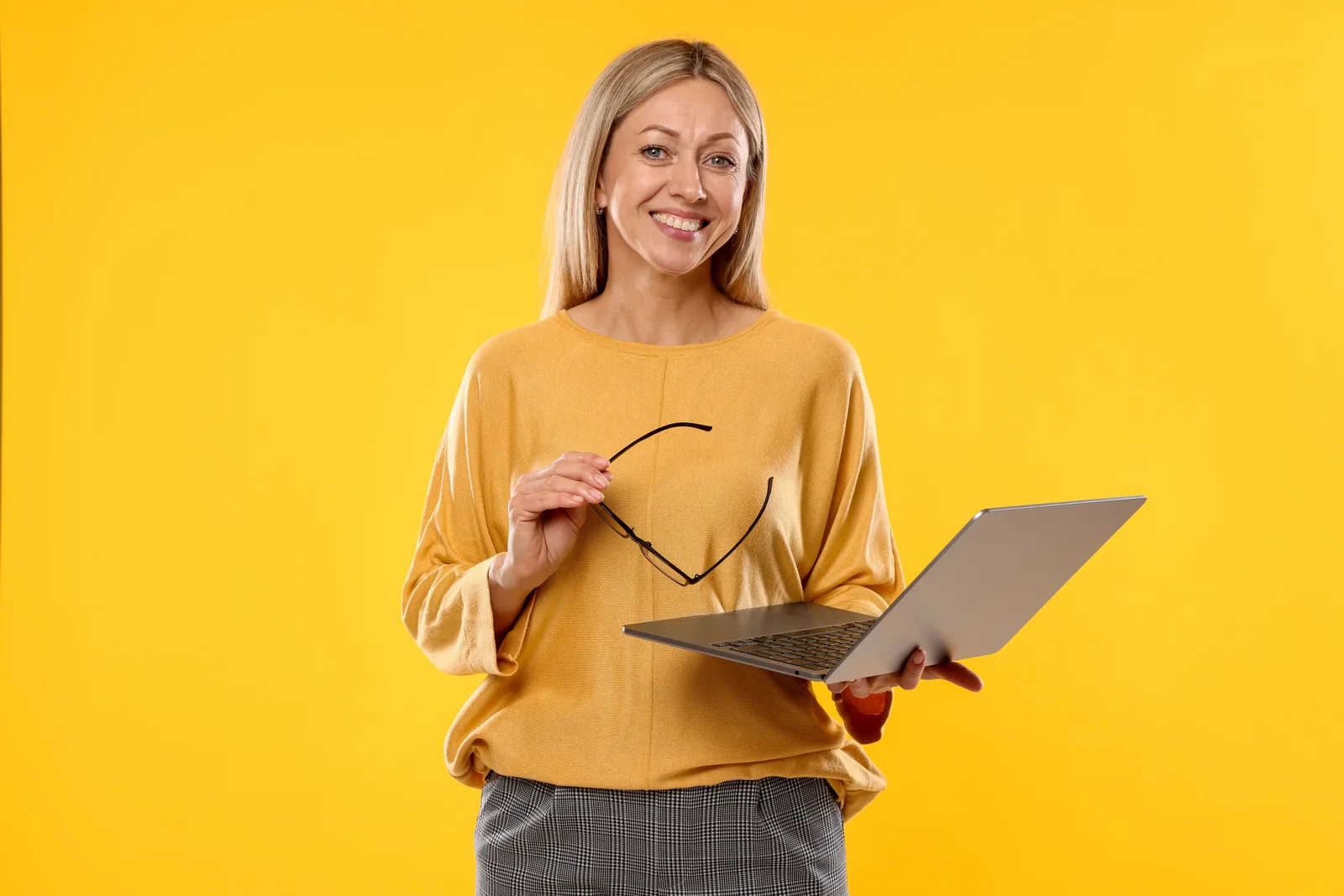 Mulher sorridente segurando um notebook aberto e os óculos, sobre fundo amarelo